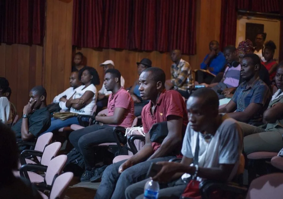 An audience in a theatre, seated in rows of pink chairs