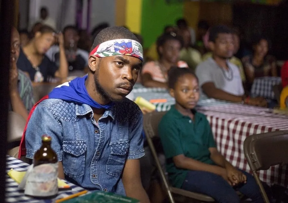 A young man in a headband and denim shirt at a table, an audience behind him