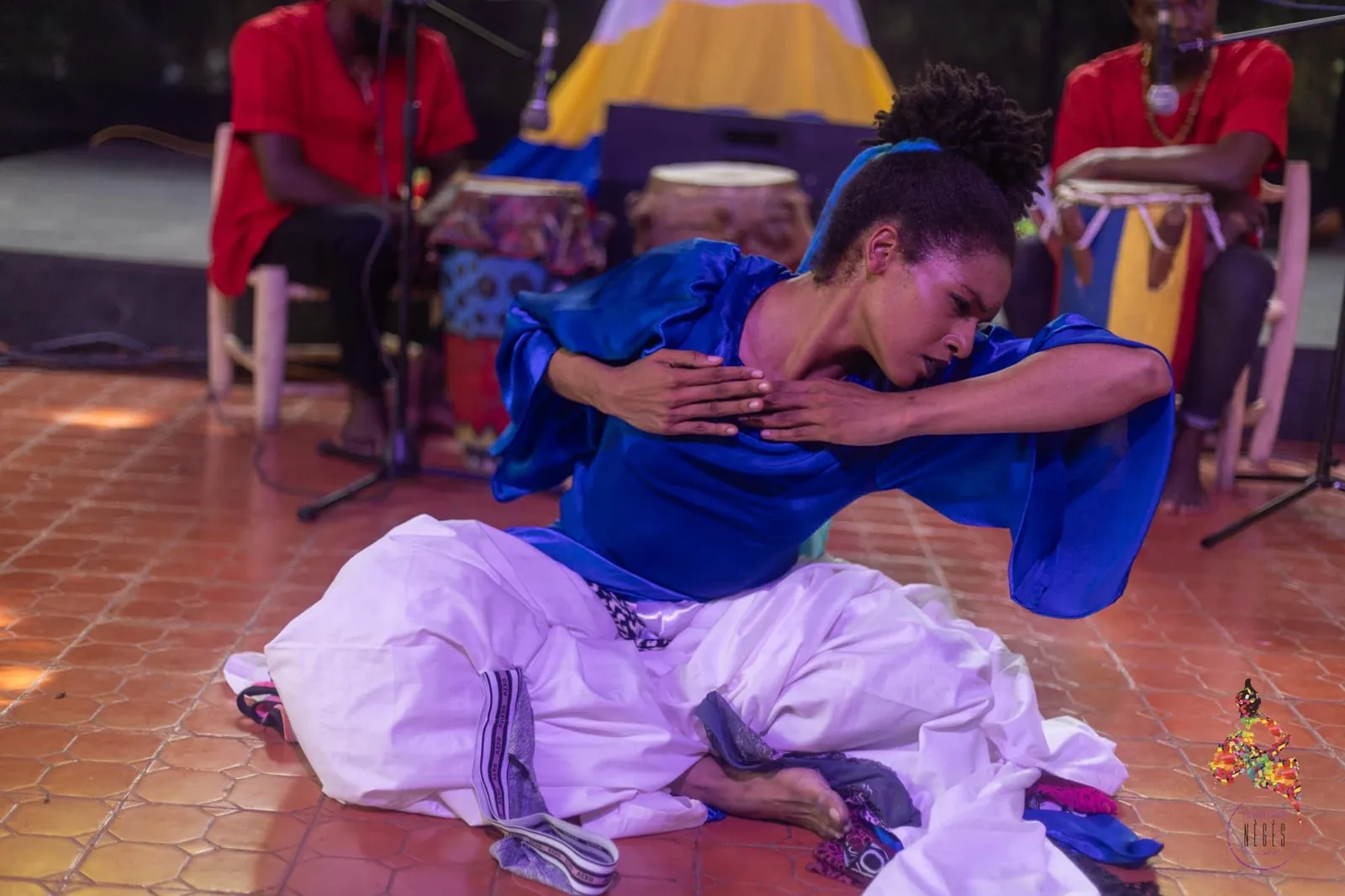 A dancer in blue and white bent low to the floor, drummers behind her